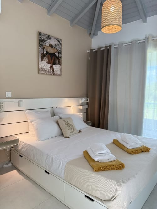 Chambre moderne à la Domaine de la Glacière avec lit blanc, luminaire suspendu tressé, œuvres tropicales et rideaux vue océan