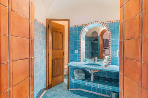 Salle de bains lumineuse aux carrelages turquoise et orange, lavabo bleu sur piédestal, miroir voûté à Villa Marina Di Cavu, Bonifacio