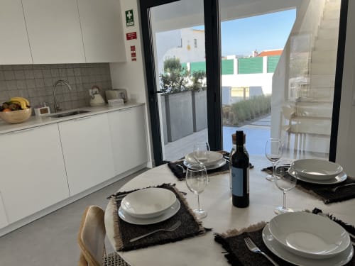 Sala de jantar de villa moderna com vista para o mar, mesa posta com taças de vinho, cozinha e acesso ao terraço exterior em Bordeira.