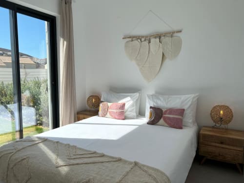 Quarto luminoso com roupa de cama branca, almofadas rosa, arte de folhas na parede e vistas para o mar na villa Casa Amoreira em Bordeira, Portugal