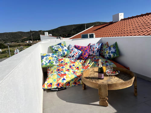 Área de estar colorida no terraço da villa Casa Arrifana em Bordeira com almofadas estampadas, mesa de madeira, vinho e