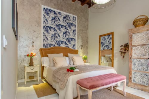 Dormitorio luminoso con cabecero beige, cuadros botánicos azules, banco rosa y cómoda de madera en apartamento de Puerto Santa María