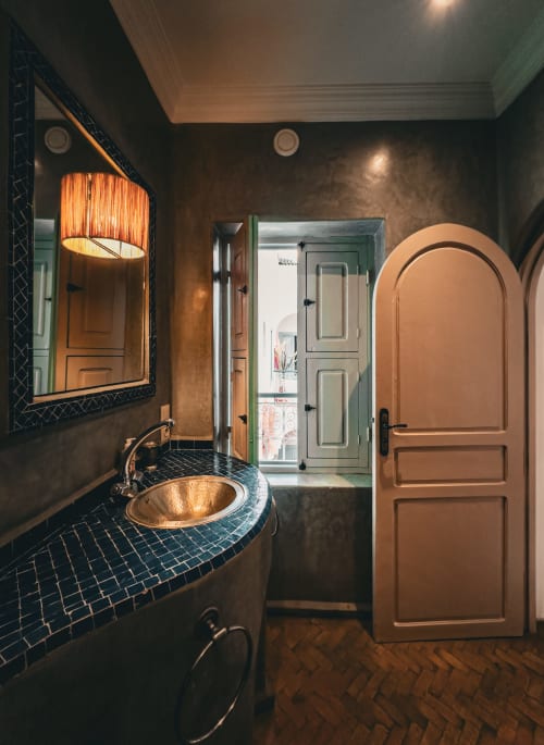 Salle de bain luxueuse au Riad Les Lauriers Blancs à Marrakech avec lavabo en cuivre, murs carrelés foncés et porte en bois arquée