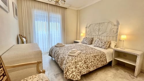 Elegant bedroom in Marbella apartment with patterned bedding, ornate headboard, balcony doors, and warm lighting