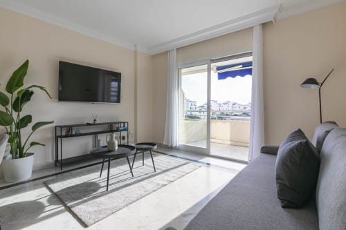Modern living room in Marina Banús apartment with sea view, gray sofa, wall-mounted TV, and balcony overlooking Marbella
