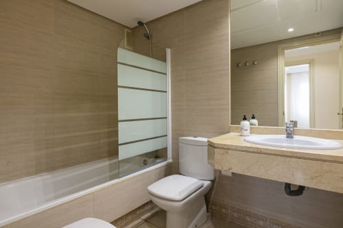 Modern bathroom in Marbella apartment with beige tile, glass shower enclosure, white vanity sink, and toilet at Marina Banús.