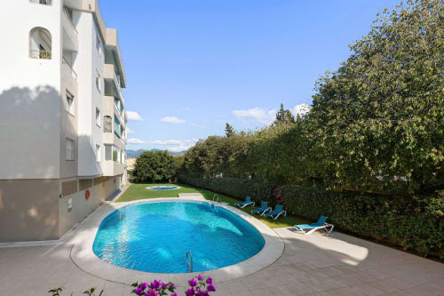 Circular pool at Marbella apartment with mountain views, lounge chairs, hedges, and white building in Nueva Andalucía.