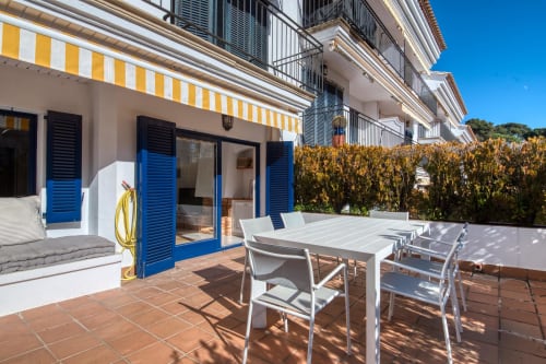 Beachfront apartment terrace in Palafrugell with white dining table, blue shutters, yellow striped awning, and direct beach