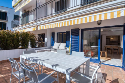 Beachfront apartment patio in Palafrugell with dining table, lounge seating, striped awning, and blue shuttered doors