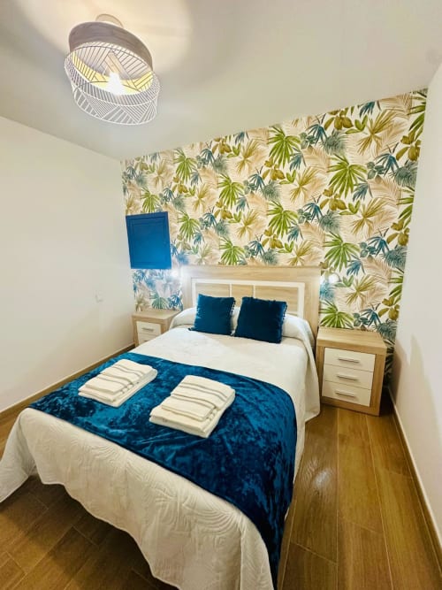 Habitación moderna en apartamento de Mérida con papel tapiz tropical, cama de madera, manta azul de terciopelo y luminaria contemporánea.