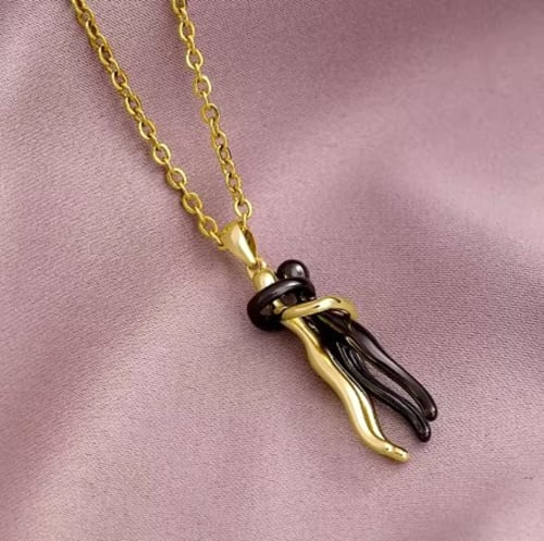 Gold chain necklace with black and gold hand pendant on pink fabric at Les Secrets de Winna