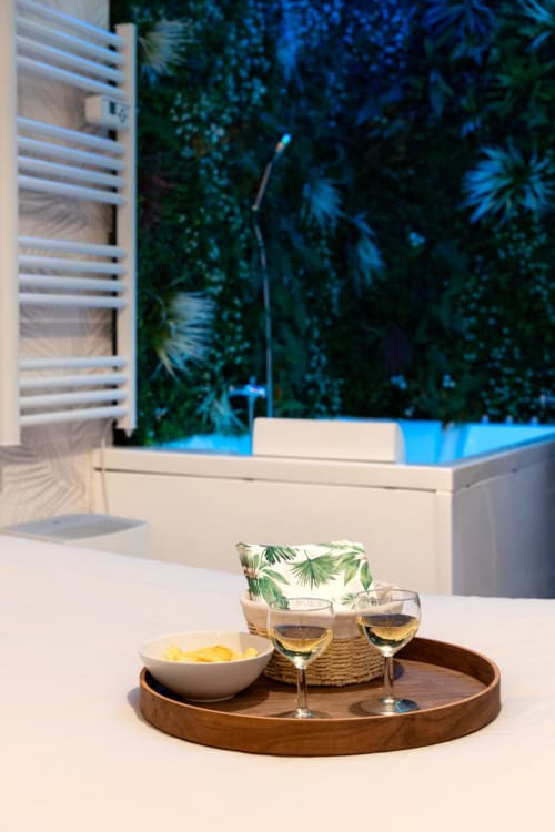 Elegant Bordeaux apartment balcony with wine glasses, bowl of chips, tropical plants, and illuminated Jacuzzi pool at dusk