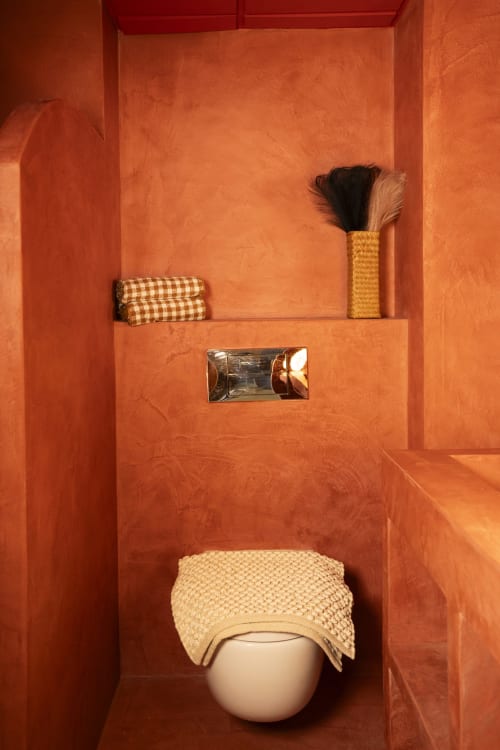 Baño naranja brillante en estudio de Formentera con inodoro blanco, accesorios tejidos, estante con toallas y cepillo