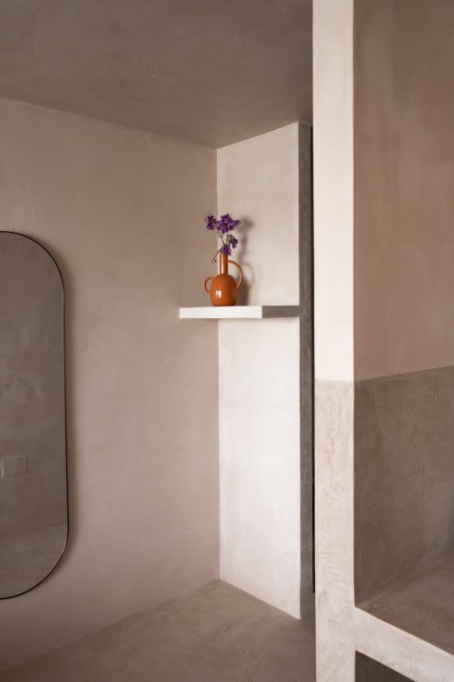 Baño minimalista en Formentera Especial con estantería blanca, jarrón cerámico naranja y flores púrpura, pared neutra