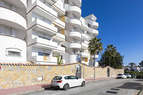 Modern white apartment building in Marbella with curved balconies, stone wall, and palm trees under clear blue sky