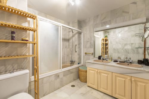 Luxurious bathroom with marble tiles, dual vanity sinks, wooden storage shelves, and glass shower enclosure in Marbella