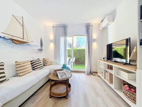 Sala de estar luminosa de la casita La Vela en Estartit con sofá blanco, cuadro de velero y vistas al jardín a través de puertas de cristal