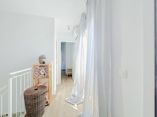 Pasillo luminoso del cottage La Vela en Estartit Rental con paredes blancas, cortinas translúcidas, suelo de madera y almacenamiento de mimbre