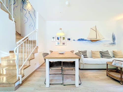 Sala de estar brillante con decoración náutica, cuadro de velero, mesa de madera, cojines a rayas y escalera de caracol
