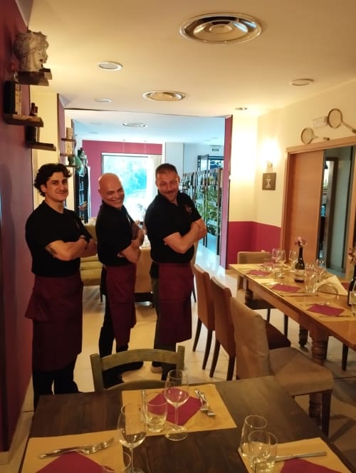 Tre addetti al ristorante in camicie nere e grembiuli bordeaux in piedi nella sala da pranzo dell'Hotel Milomax con tavoli preparati