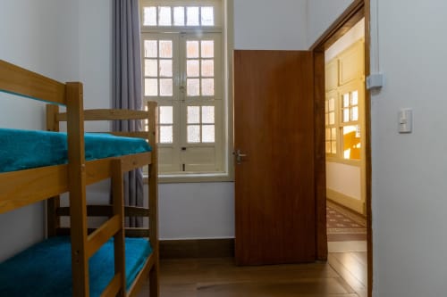Beliche de madeira com colchões azul-petróleo no quarto duplo do albergue Rio Vibes, com grande janela e porta de madeira marrom para áreas compartilhadas