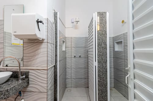 Banheiro compartilhado moderno no Rio Vibes Hostel com chuveiro aberto, pia de granito e prateleiras brancas