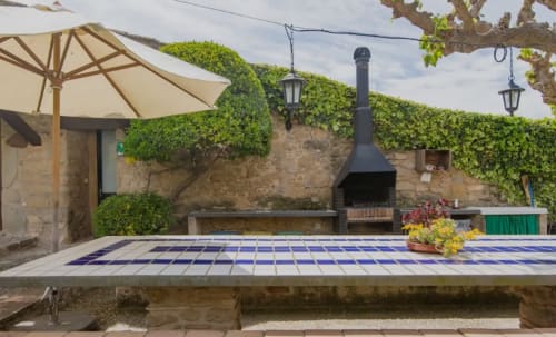 Patio de Can Gat Vell con mesa de azulejos azules, chimenea de piedra, muros cubiertos de hiedra y sombrilla en Empordà, Girona