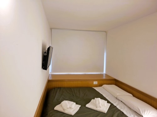 Camera da letto compatta di Newhouse Bari con letto in legno, biancheria verde, asciugamani arrotolati, TV a muro