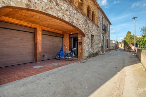 Maison en pierre avec portes de garage en bois et scooter bleu garé sous carport, Palafrugell