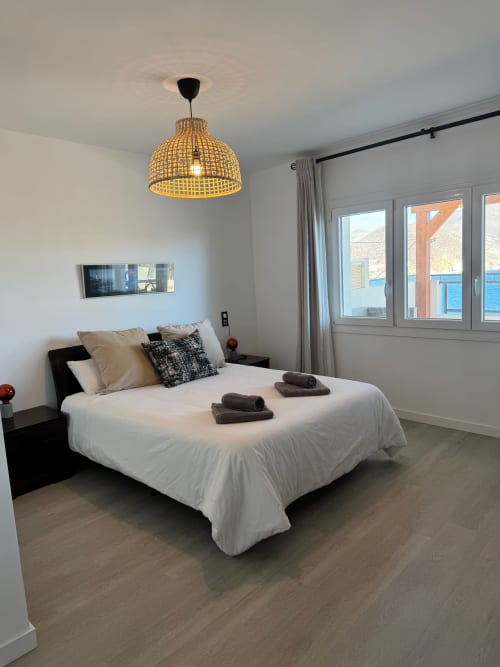 Dormitorio moderno en El Faro De Los Genoveses con vista al mar, cama blanca, lámpara colgante amarilla y acceso a balcón costero