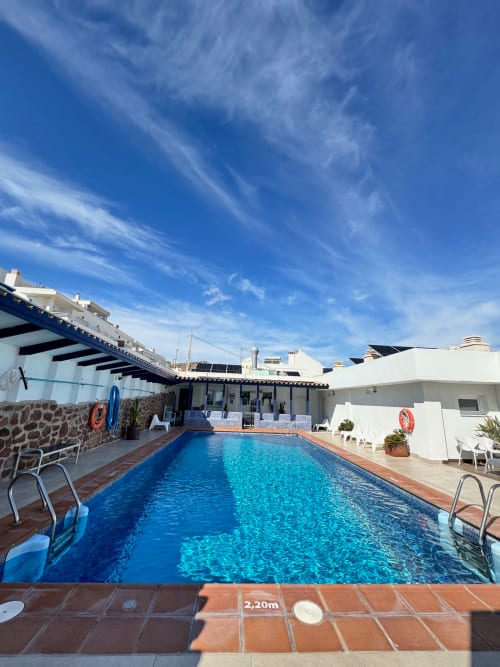 Piscina rectangular del hotel El Faro De Los Genoveses en San José, Almería, con cielo azul y edificios costeros