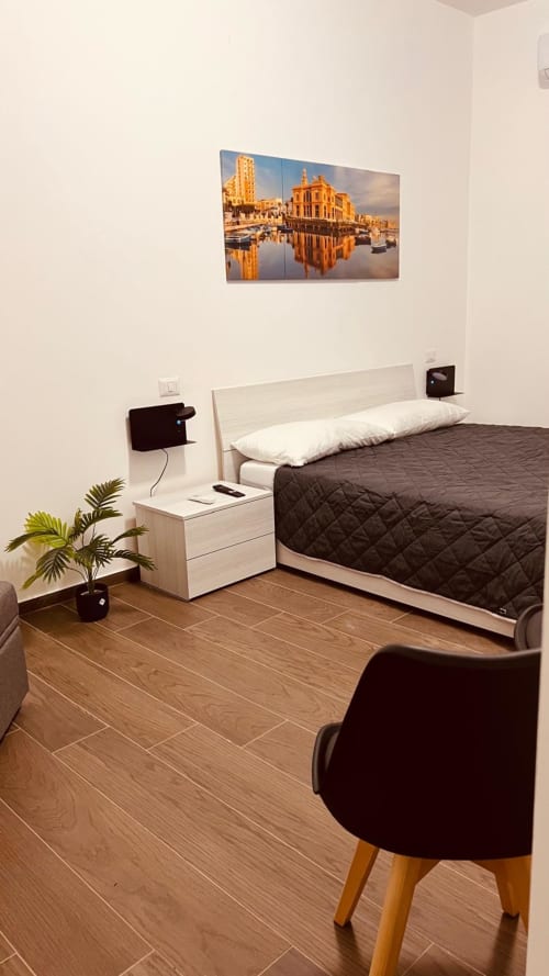 Camera da letto moderna a Bari Vacation con pavimento in legno, letto bianco, biancheria scura, comodino, poltrona, pianta in vaso e vista sulla città