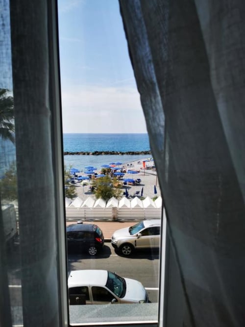 Vista dalla finestra dell'appartamento di TuffonelBlu, Ventimiglia: spiaggia con ombrelloni blu, parcheggio e Mar Mediterraneo