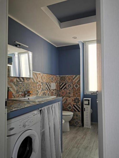 Moderno bagno con pareti blu navy, piastrelle decorate, lavabo, wc, lavatrice e finestra a TuffonelBlu