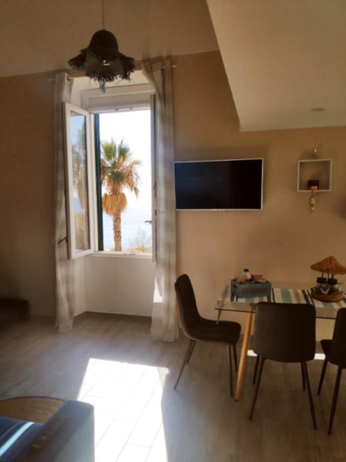 Luminoso appartamento moderno a Ventimiglia con vista mare, tavolo da pranzo, TV e palme visibili dalla porta aperta