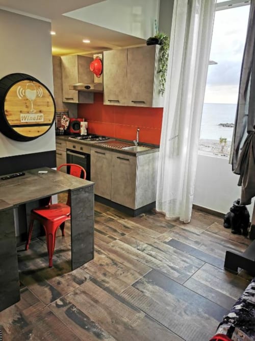 Moderna cucina con rivestimento rosso, zona pranzo e vista mare dalle porte della veranda nell'appartamento di Ventimiglia