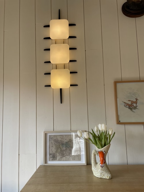 Modern wall sconce with three frosted glass shades above wooden shelf at Clairseine cottage, with white tulips in decorative