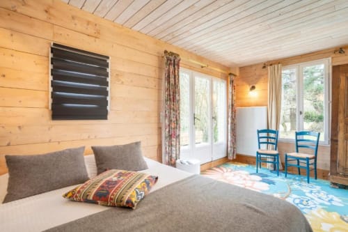 Bright wooden cottage bedroom with garden views, flat-screen TV, blue chairs, and colorful rug at Les logis de Kergoulou,