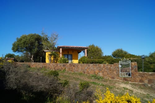 Yellow cottage villa with stone wall and white gate in Orosei, surrounded by Mediterranean vegetation and clear blue sky