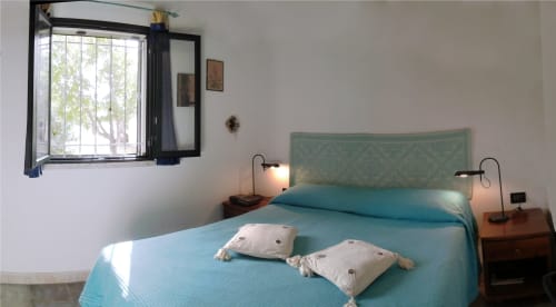 Bright bedroom with turquoise bed, two decorative pillows, wooden nightstands with lamps, and barred window overlooking