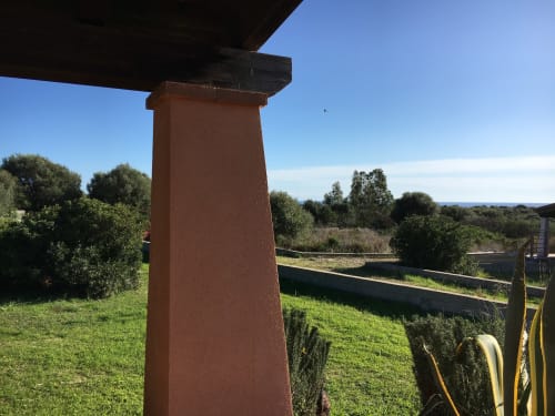 Rusty red metal support beam under pergola with green lawn, shrubs, and distant sea view at Orosei villa