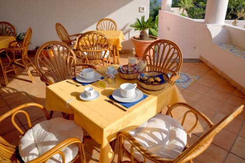 Soleggiata zona colazione dell'Hotel Principe di Rinella con sedie in vimini, tavolo giallo, piante in vaso e stoviglie bianche