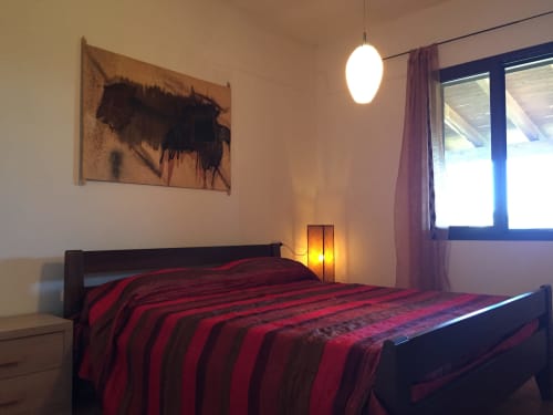 Modern bedroom with red striped bedding, dark bed frame, pendant light, abstract wall art, and blue-framed window in Orosei