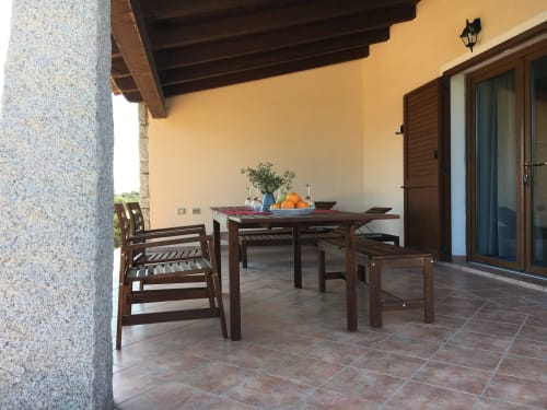 Wooden dining table and chairs on a terracotta tiled veranda with yellow walls at Domo Sara villa in Orosei, Italy