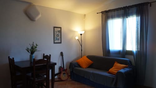 Cozy Orosei villa living room with black sofa, wooden dining table, guitar, and sea views through sheer curtains