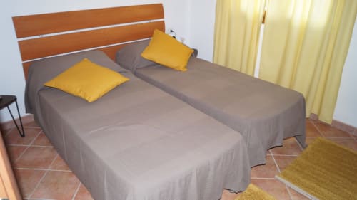 Twin beds with gray linens and yellow pillows in Domo Sara villa bedroom, Orosei, with terracotta tiles and wooden headboard.
