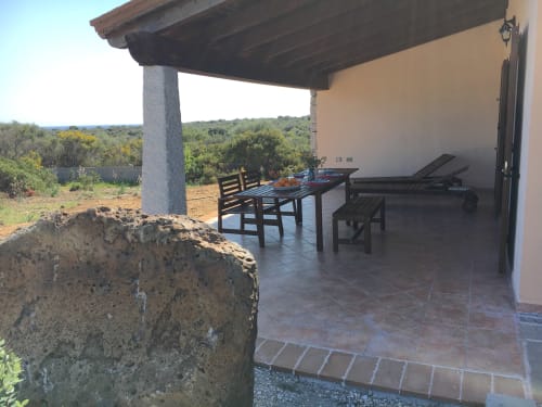 Covered veranda of Domo Sara villa in Orosei with wooden furniture, dining table, and sea view over Mediterranean landscape