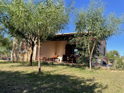 Tuscan villa in Orosei with shaded veranda, wooden furnishings, and lush garden under clear sky