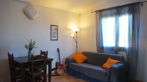 Cozy living room in Domo Sara villa, Orosei with gray sofa, wooden dining table, guitar, and bright windows