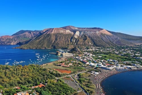 Vista aerea dell'Hotel Principe di Rinella a Rinella, Sicilia, con vulcano, baia turchese e villaggio costiero